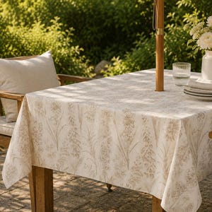 Può includere: Una tovaglia rettangolare beige con un motivo floreale copre un tavolo di legno. Il tavolo è apparecchiato all'aperto con un ombrellone di legno, un bicchiere d'acqua e un vaso di fiori bianchi. È visibile una sedia di legno con un cuscino beige.