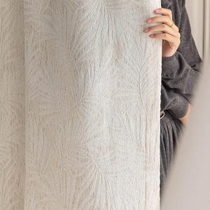 Könnte beinhalten: Cremefarbener Vorhang mit einem dezenten, sich wiederholenden Blattmuster. Der Stoff hat ein strukturiertes Aussehen, und das Design ist über die gesamte Oberfläche sichtbar. Der Vorhang wird von einer Person in einem grauen Pullover gehalten.