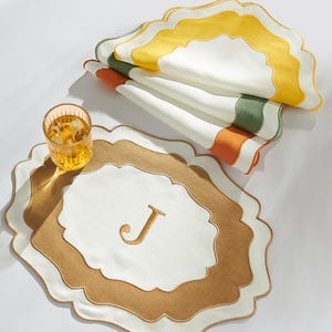 May include: A stack of scalloped-edge placemats in white with yellow, green, and orange stripes. A matching placemat with a gold embroidered "J" is in the foreground, next to a glass of iced beverage. The placemats are likely made of fabric.
