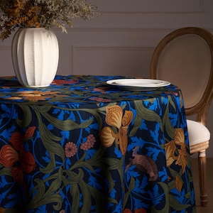 May include: A round tablecloth featuring a dark blue background with a floral design in orange, green, and blue. A white vase with dried flowers and a white plate are on the table. A wooden chair is in the background.