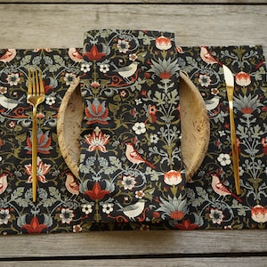 May include: A rectangular placemat and a folded napkin featuring a dark background with a repeating pattern of birds, red flowers, and green foliage. A gold-toned fork is placed on the left side, a gold-toned knife on the right, and a wooden bowl.