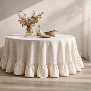 Puede incluir: Un mantel redondo de lino blanco roto con borde con volantes. La mesa está puesta con un jarrón de flores secas, cristalería, una botella, un cuenco y un plato. El mantel tiene aproximadamente 183 cm de diámetro.