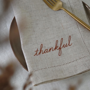 May include: A light beige linen napkin featuring the word thankful embroidered in rust-coloured thread. The napkin has a decorative hemstitch border and is placed on a plate with a gold-toned metal fork resting on top of the fabric surface.