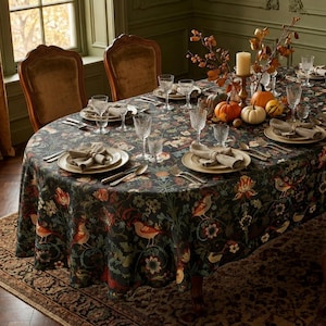 May include: A dark-colored tablecloth featuring a repeating pattern of birds, flowers, and foliage in shades of orange, green, and cream. The table is set with white plates, folded napkins, clear glassware, silverware, and decorative pumpkins.