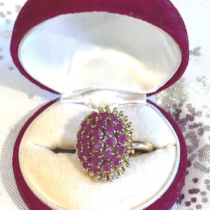 May include: A gold ring with a cluster of small, round, deep pink gemstones. The ring is displayed in an open, burgundy velvet ring box with a white satin interior. The ring's design is a prominent feature.