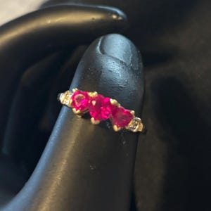May include: A gold ring featuring three heart-shaped, vibrant red gemstones. The ring has small, clear accent stones and is set on a gold band. A piece of jewelry with a romantic theme.