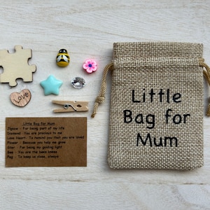 May include: A tan burlap drawstring bag with the text Little Bag for Mum printed in black. Beside it are small items: a wooden puzzle piece, a bee charm, a pink flower, a blue star, a clear gem, a wooden heart with Love, and a wooden clothespin.