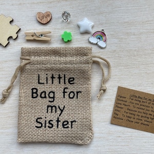 Może przedstawiać: Mały woreczek z juty z nadrukiem "Little Bag for my Sister". Woreczek jest wypełniony małymi prezentami, w tym kawałkiem układanki, diamentem, sercem, koniczyną, gwiazdą, tęczą i kołkiem.