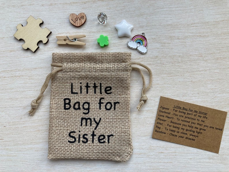 Może przedstawiać: Mały woreczek z juty z nadrukiem "Little Bag for my Sister". Woreczek jest wypełniony małymi prezentami, w tym kawałkiem układanki, diamentem, sercem, koniczyną, gwiazdą, tęczą i kołkiem.