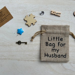 May include: A small burlap bag with the text "Little Bag for my Husband" printed on it. The bag is surrounded by various small objects, including a blue star, a key, a wooden puzzle piece, a wooden heart with the word "Love" on it, a small wooden clothespin, and a clear rhinestone.