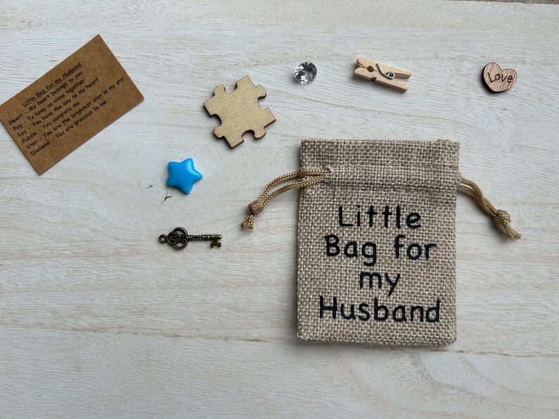 May include: A small burlap bag with the text "Little Bag for my Husband" printed on it. The bag is surrounded by various small objects, including a blue star, a key, a wooden puzzle piece, a wooden heart with the word "Love" on it, a small wooden clothespin, and a clear rhinestone.