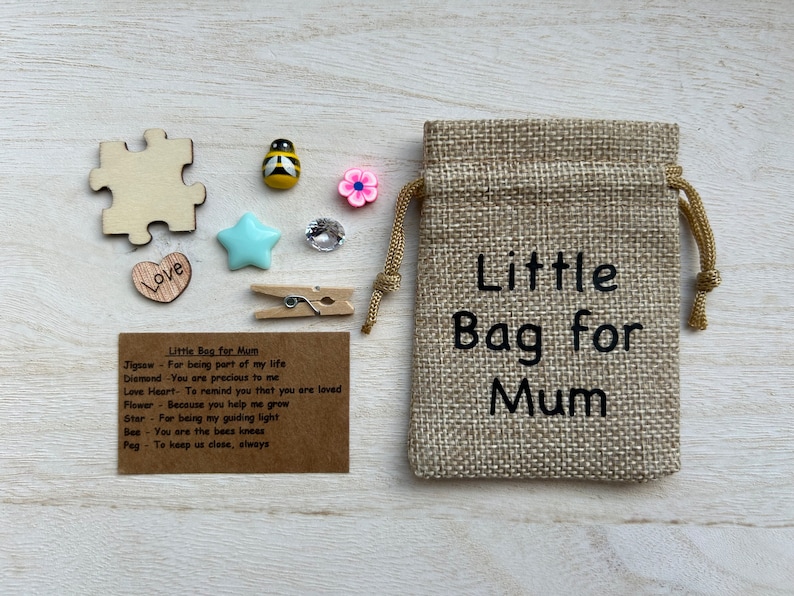 May include: A tan burlap drawstring bag with the text Little Bag for Mum printed in black. Beside it are small items: a wooden puzzle piece, a bee charm, a pink flower, a blue star, a clear gem, a wooden heart with Love, and a wooden clothespin.