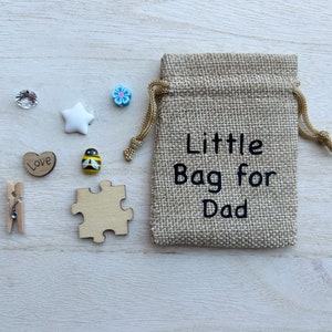 May include: A small hessian bag with the text "Little Bag for Dad" printed on it. The bag is surrounded by small decorative items, including a white star, a blue flower, a heart with the word "Love" on it, a bee, a jigsaw piece, and two clear rhinestones.