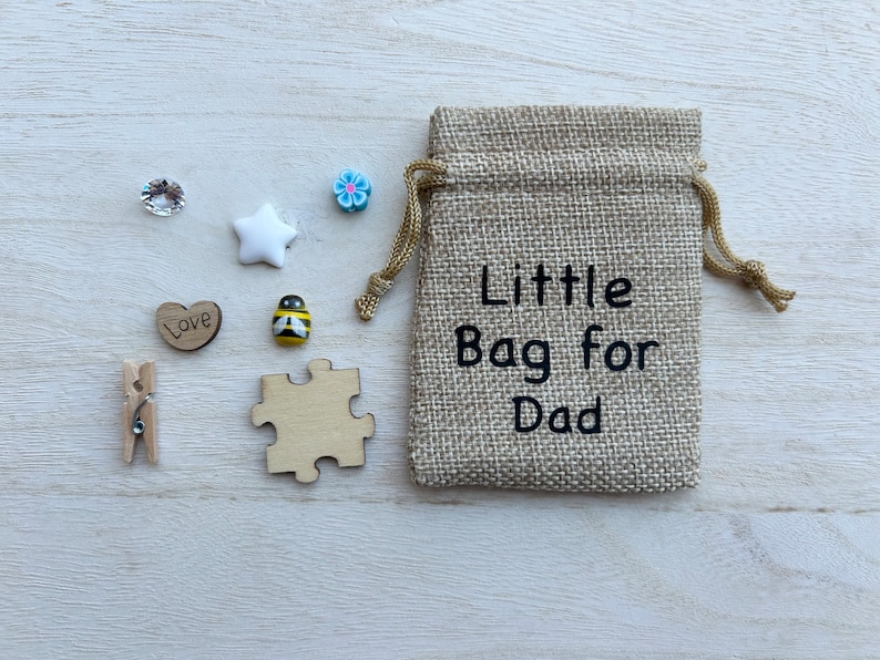 May include: A small hessian bag with the text "Little Bag for Dad" printed on it. The bag is surrounded by small decorative items, including a white star, a blue flower, a heart with the word "Love" on it, a bee, a jigsaw piece, and two clear rhinestones.