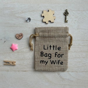 May include: A small burlap bag with a drawstring closure. The bag has the text "Little Bag for my Wife" printed on it. The bag is surrounded by small decorative items, including a wooden heart with the word "Love" on it, a pink star, a wooden puzzle piece, a small clear crystal, and a gold-colored key.