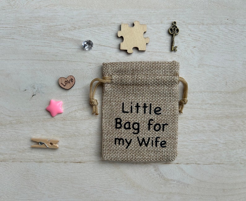May include: A small burlap bag with a drawstring closure. The bag has the text "Little Bag for my Wife" printed on it. The bag is surrounded by small decorative items, including a wooden heart with the word "Love" on it, a pink star, a wooden puzzle piece, a small clear crystal, and a gold-colored key.