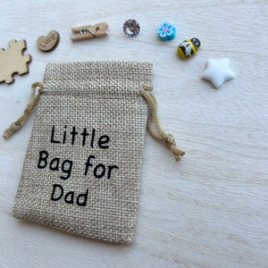 May include: A small hessian bag with a drawstring closure. The bag is decorated with the text "Little Bag for Dad". The bag is surrounded by various small decorative items, including a wooden jigsaw piece, a heart with the word "Love" on it, a wooden peg, a blue flower, a bee, and a white star.