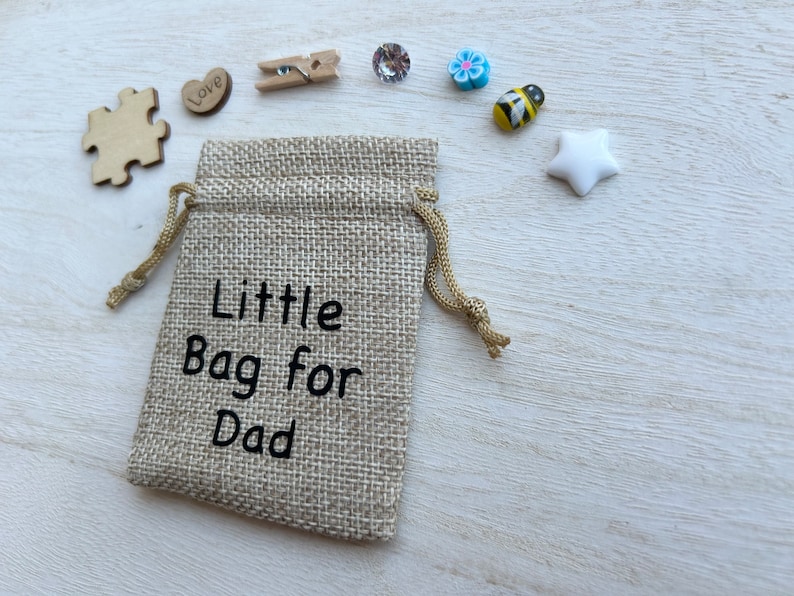 May include: A small hessian bag with a drawstring closure. The bag is decorated with the text "Little Bag for Dad". The bag is surrounded by various small decorative items, including a wooden jigsaw piece, a heart with the word "Love" on it, a wooden peg, a blue flower, a bee, and a white star.