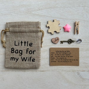 May include: A small burlap bag with the text "Little Bag for my Wife" printed on it. The bag is filled with small items, including a heart, a key, a star, a puzzle piece, a peg, and a diamond. A card with the text "Little Bag for my Wife" and descriptions of each item is included.