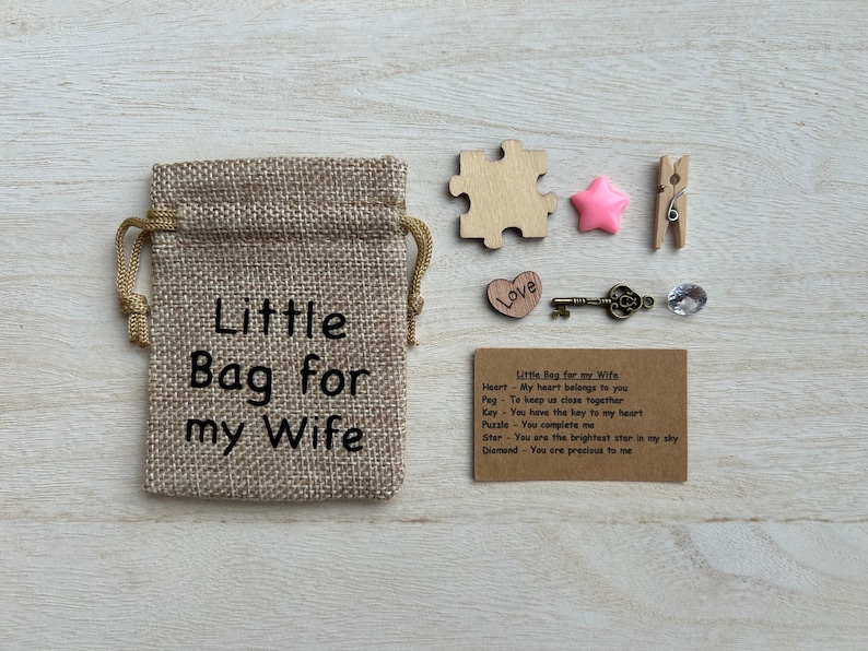 May include: A small burlap bag with the text "Little Bag for my Wife" printed on it. The bag is filled with small items, including a heart, a key, a star, a puzzle piece, a peg, and a diamond. A card with the text "Little Bag for my Wife" and descriptions of each item is included.