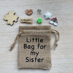 Może przedstawiać: Mały woreczek z juty ze sznurkiem do ściągania. Woreczek jest ozdobiony napisem "Little Bag for my Sister". Woreczek jest otoczony r&oacute;żnymi małymi przedmiotami, w tym drewnianym elementem układanki, drewnianym sercem z napisem "Love", małą zieloną koniczyną, białą gwiazdą, przezroczystym kryształem i zawieszką w kształcie tęczy.