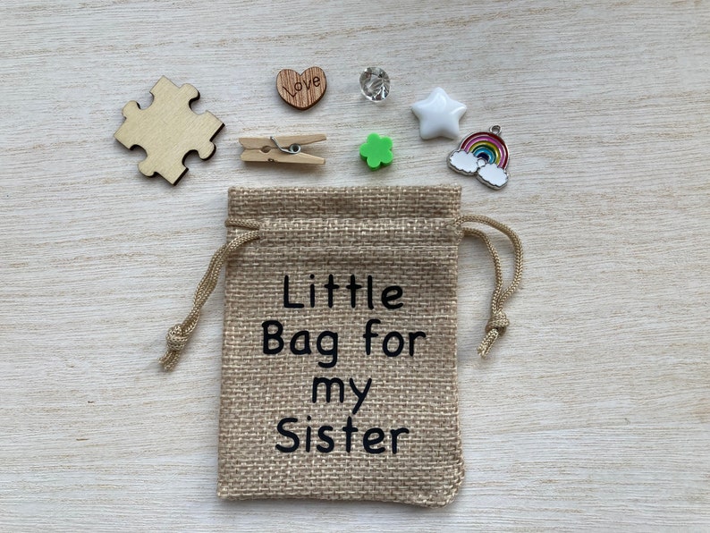 Może przedstawiać: Mały woreczek z juty ze sznurkiem do ściągania. Woreczek jest ozdobiony napisem "Little Bag for my Sister". Woreczek jest otoczony r&oacute;żnymi małymi przedmiotami, w tym drewnianym elementem układanki, drewnianym sercem z napisem "Love", małą zieloną koniczyną, białą gwiazdą, przezroczystym kryształem i zawieszką w kształcie tęczy.