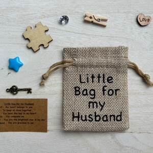 May include: A small burlap bag with the text "Little Bag for my Husband" printed on it. The bag is surrounded by various small objects, including a heart, a star, a key, a puzzle piece, and a diamond.