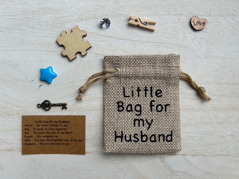 May include: A small burlap bag with the text "Little Bag for my Husband" printed on it. The bag is surrounded by various small objects, including a heart, a star, a key, a puzzle piece, and a diamond.