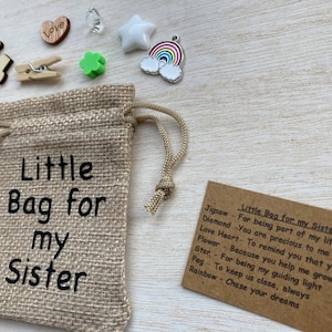 Może przedstawiać: Mały woreczek z juty z nadrukiem "Little Bag for my Sister". Woreczek jest wypełniony małymi ozdobami, w tym kawałkiem układanki, sercem, koniczyną, gwiazdą, wisiorkiem w kształcie tęczy i kołkiem. Obok woreczka znajduje się karta z tekstem "Little Bag for my Sister" i listą przedmiot&oacute;w oraz ich znaczeń.