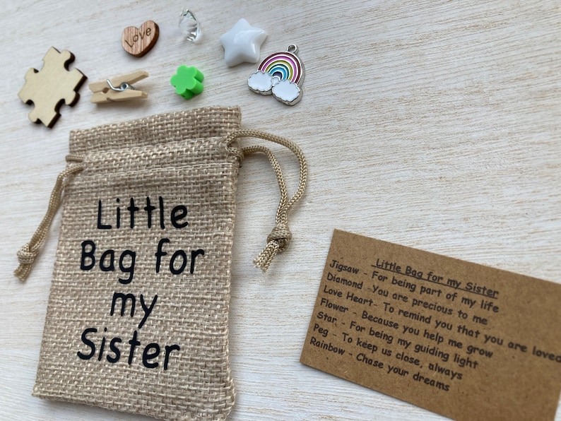 Może przedstawiać: Mały woreczek z juty z nadrukiem "Little Bag for my Sister". Woreczek jest wypełniony małymi ozdobami, w tym kawałkiem układanki, sercem, koniczyną, gwiazdą, wisiorkiem w kształcie tęczy i kołkiem. Obok woreczka znajduje się karta z tekstem "Little Bag for my Sister" i listą przedmiot&oacute;w oraz ich znaczeń.