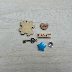 May include: A wooden puzzle piece, a wooden heart with the word "Love" written on it, a wooden clothespin, a small blue star, a clear rhinestone, and a small antique-style key on a white wooden background.