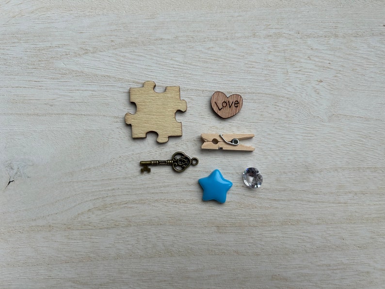 May include: A wooden puzzle piece, a wooden heart with the word "Love" written on it, a wooden clothespin, a small blue star, a clear rhinestone, and a small antique-style key on a white wooden background.
