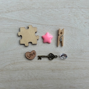 May include: A wooden puzzle piece, a pink star, a wooden clothespin, a wooden heart with the word "Love" engraved on it, a key, and a clear crystal on a light wood background.