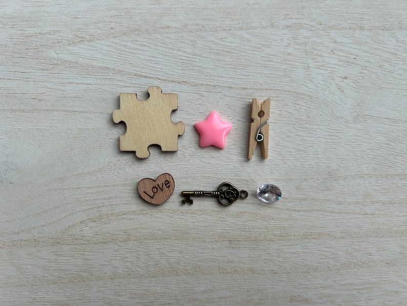 May include: A wooden puzzle piece, a pink star, a wooden clothespin, a wooden heart with the word "Love" engraved on it, a key, and a clear crystal on a light wood background.