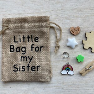Może przedstawiać: Mały woreczek z juty z nadrukiem "Little Bag for my Sister". Woreczek jest wypełniony małymi ozdobami, w tym drewnianym sercem z napisem "Love", białą gwiazdą, przezroczystym kryształem, zieloną koniczyną, wisiorkiem w kształcie tęczy i drewnianym klips.