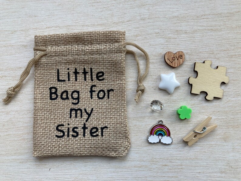 Może przedstawiać: Mały woreczek z juty z nadrukiem "Little Bag for my Sister". Woreczek jest wypełniony małymi ozdobami, w tym drewnianym sercem z napisem "Love", białą gwiazdą, przezroczystym kryształem, zieloną koniczyną, wisiorkiem w kształcie tęczy i drewnianym klips.