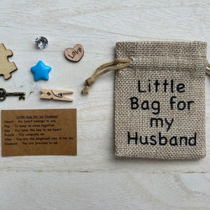 May include: A small burlap bag with a drawstring closure. The bag is decorated with the text "Little Bag for my Husband". The bag is filled with small items, including a heart, a star, a key, a peg, a puzzle piece, and a diamond. A card with the text "Little Bag for my Husband" and a list of items with their meanings is included.