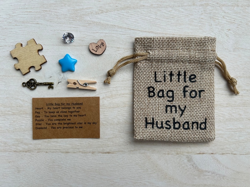 May include: A small burlap bag with a drawstring closure. The bag is decorated with the text "Little Bag for my Husband". The bag is filled with small items, including a heart, a star, a key, a peg, a puzzle piece, and a diamond. A card with the text "Little Bag for my Husband" and a list of items with their meanings is included.