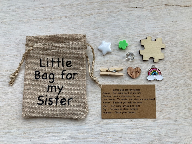 Może przedstawiać: Mały woreczek z juty z nadrukowanym tekstem "Little Bag for my Sister". Woreczek zawiera r&oacute;żne małe przedmioty, w tym drewniany element układanki, diamentowy wisiorek, wisiorek w kształcie serca, wisiorek w kształcie kwiatu, wisiorek w kształcie gwiazdy, kołek i wisiorek w kształcie tęczy. Dołączona jest karta z tekstem "Little Bag for my Sister" i listą znaczeń dla każdego przedmiotu.