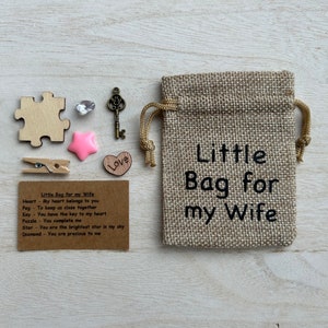 May include: A small burlap bag with the text "Little Bag for my Wife" printed on it. The bag contains a variety of small items, including a wooden heart, a wooden puzzle piece, a wooden peg, a key, a star, and a diamond. A card with the text "Little Bag for my Wife" and a list of meanings for each item is included.