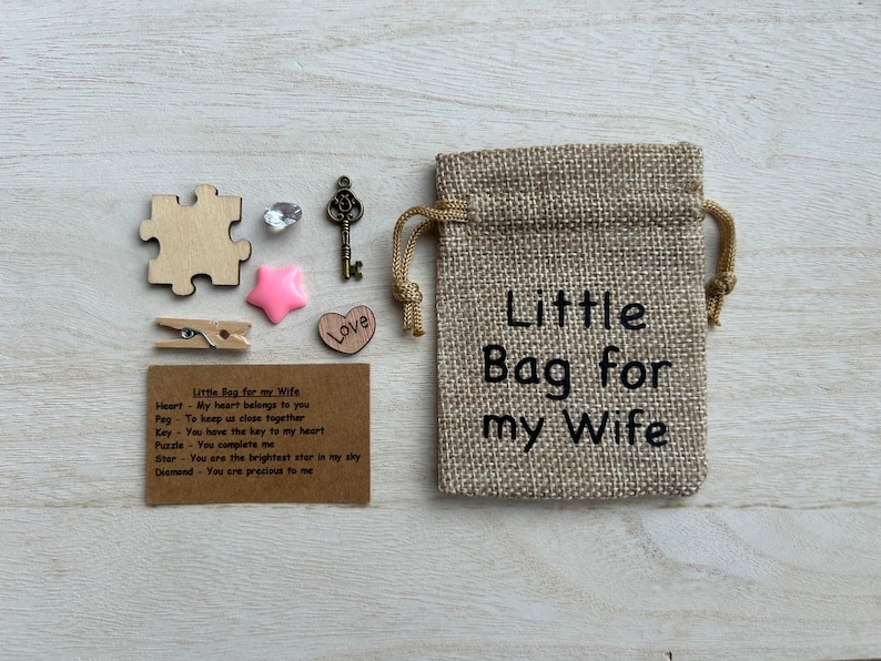 May include: A small burlap bag with the text "Little Bag for my Wife" printed on it. The bag contains a variety of small items, including a wooden heart, a wooden puzzle piece, a wooden peg, a key, a star, and a diamond. A card with the text "Little Bag for my Wife" and a list of meanings for each item is included.