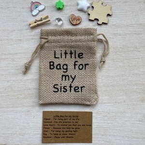 Może przedstawiać: Mały woreczek z juty z nadrukiem "Little Bag for my Sister". Woreczek jest wypełniony małymi ozdobami, w tym kawałkiem układanki, sercem, gwiazdą, kwiatem, kołkiem i tęczą. Dołączona jest karta z tekstem "Little Bag for my Sister" i listą znaczeń dla każdej ozdoby.