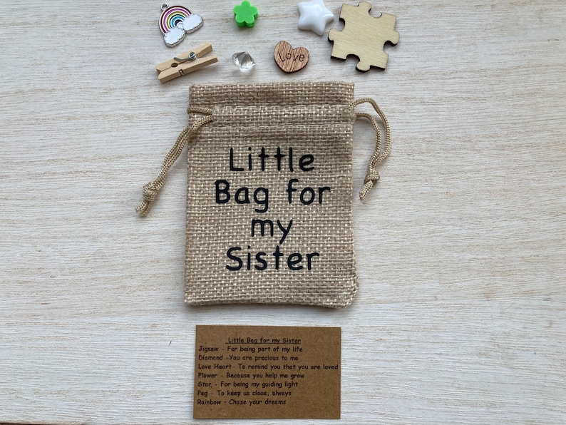Może przedstawiać: Mały woreczek z juty z nadrukiem "Little Bag for my Sister". Woreczek jest wypełniony małymi ozdobami, w tym kawałkiem układanki, sercem, gwiazdą, kwiatem, kołkiem i tęczą. Dołączona jest karta z tekstem "Little Bag for my Sister" i listą znaczeń dla każdej ozdoby.