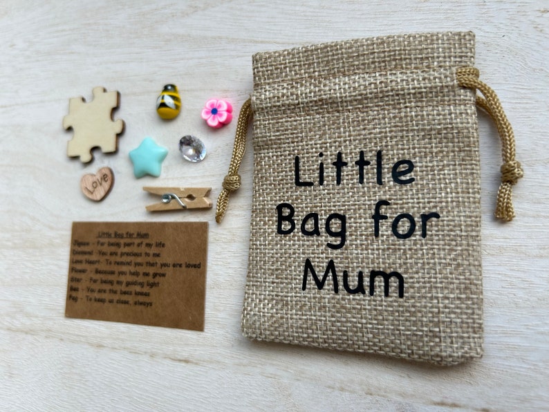 Może przedstawiać: Mały woreczek jutowy ze sznurkiem do ściągania. Woreczek jest nadrukowany tekstem "Little Bag for Mum". Woreczek jest otoczony r&oacute;żnymi małymi przedmiotami, w tym drewnianym elementem układanki, sercem, gwiazdą, pszczołą, kwiatem, spinaczem do bielizny i kartką z tekstem "Little Bag for Mum".