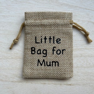 Może przedstawiać: Mały woreczek jutowy ze sznurkiem do ściągania. Woreczek jest beżowy i ma wyszyty na przodzie czarny napis "Little Bag for Mum".