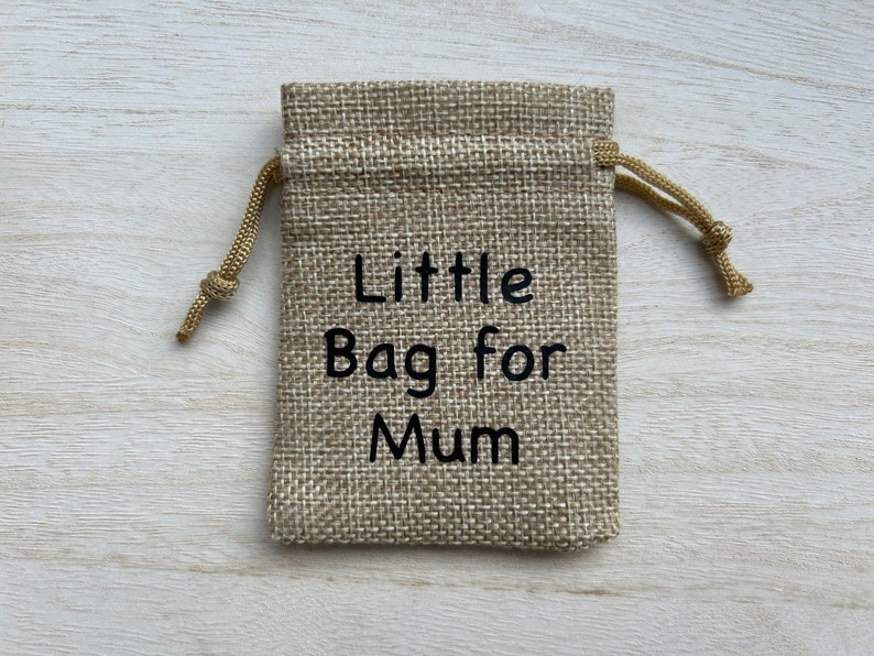 Może przedstawiać: Mały woreczek jutowy ze sznurkiem do ściągania. Woreczek jest beżowy i ma wyszyty na przodzie czarny napis "Little Bag for Mum".