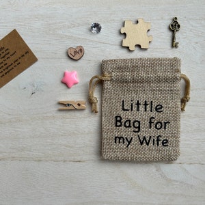 May include: A small burlap bag with the text "Little Bag for my Wife" printed on it. The bag is surrounded by various small items, including a heart, a star, a key, a puzzle piece, and a note with the text "Heart - You have the key to my heart, Pag - To keep us close together, Key - You are the key to my heart, Puzzle - You complete me, Star - You are the brightest star in my sky, Diamond - You are precious to me".