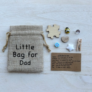 May include: A small burlap bag with the text "Little Bag for Dad" printed on it. The bag contains a variety of small items, including a wooden jigsaw piece, a wooden heart with the word "Love" on it, a small blue flower, a white star, a small wooden peg, a clear crystal, and a small wooden bee with black and yellow stripes. A card with the text "Little Bag for Dad" and a list of items with their meanings is also included.