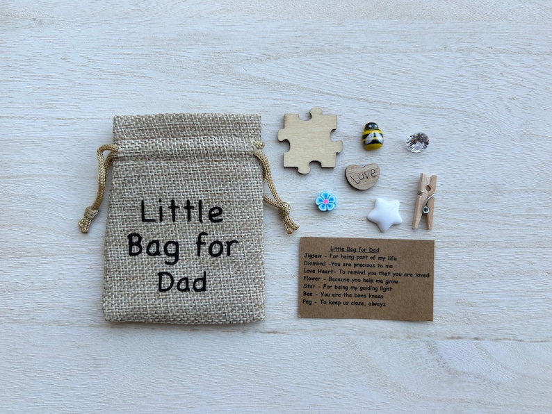 May include: A small burlap bag with the text "Little Bag for Dad" printed on it. The bag contains a variety of small items, including a wooden jigsaw piece, a wooden heart with the word "Love" on it, a small blue flower, a white star, a small wooden peg, a clear crystal, and a small wooden bee with black and yellow stripes. A card with the text "Little Bag for Dad" and a list of items with their meanings is also included.