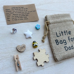 May include: A small hessian bag with the text "Little Bag for Dad" printed on it. The bag is filled with small trinkets, including a wooden heart with the word "Love" on it, a wooden jigsaw puzzle piece, a wooden peg, a white star, a blue flower, a clear crystal, and a yellow and black bee.